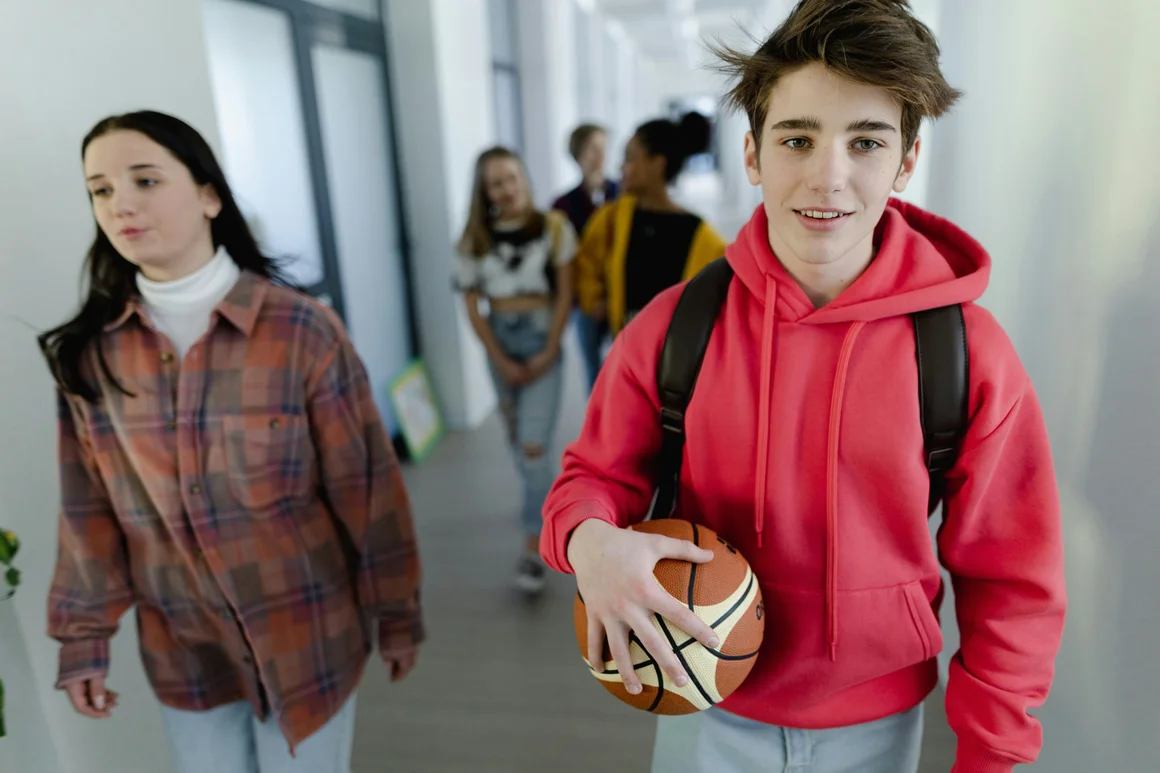 Ein Junge geht in der Schule mit einem Basketball in der Hand durch den Gang, neben und hinter ihm sind andere Schülerinnen und Schüler zu sehen