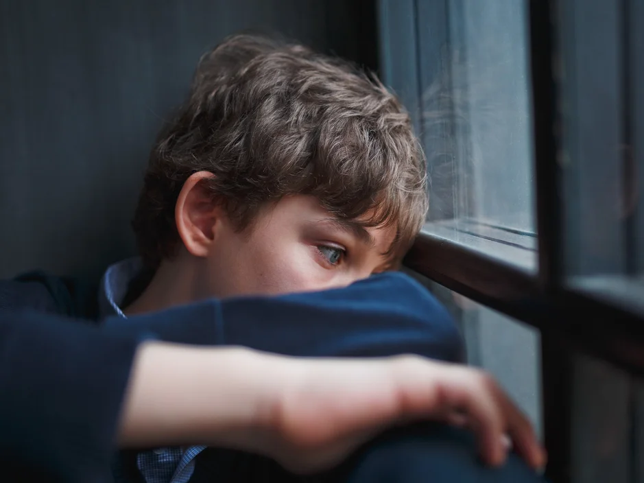Nachdenklicher Teenager sitzt am Fenster und bedeckt sein Gesicht mit den Händen